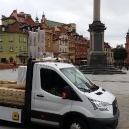 Biała furgonetka z ładunkiem tarcicy budowlanej na tle Starego Miasta w Warszawie, widoczna kolumna Zygmunta i Zamek Królewski.