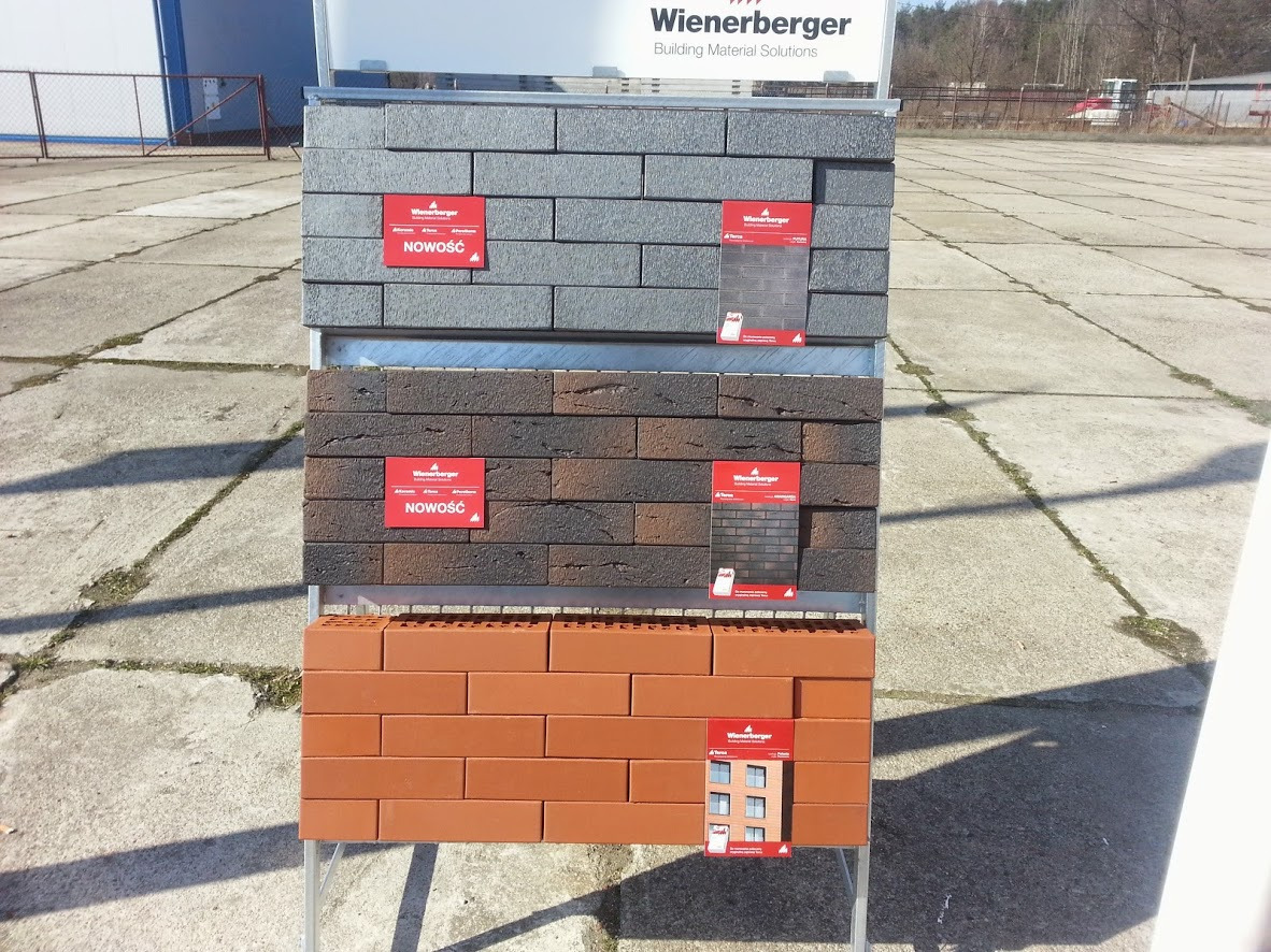 Ekspozycja trzech rodzajów cegieł elewacyjnych Wienerberger w odcieniach szarości, brązu i czerwieni z etykietami 'Nowość' na szarym betonowym podłożu.