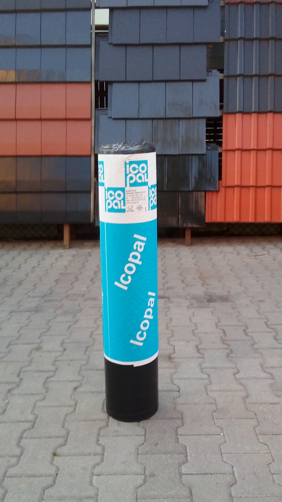 Rolka papy termozgrzewalnej marki Icopal na tle ekspozycji różnokolorowych dachówek ceramicznych.