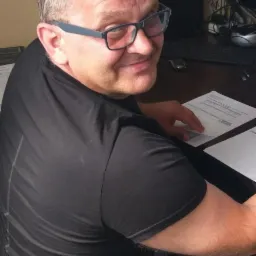 Uśmiechnięty mężczyzna w okularach siedzi przy biurku z dokumentami, spoglądając przez ramię w stronę kamery. W tle monitor i tablica informacyjna.