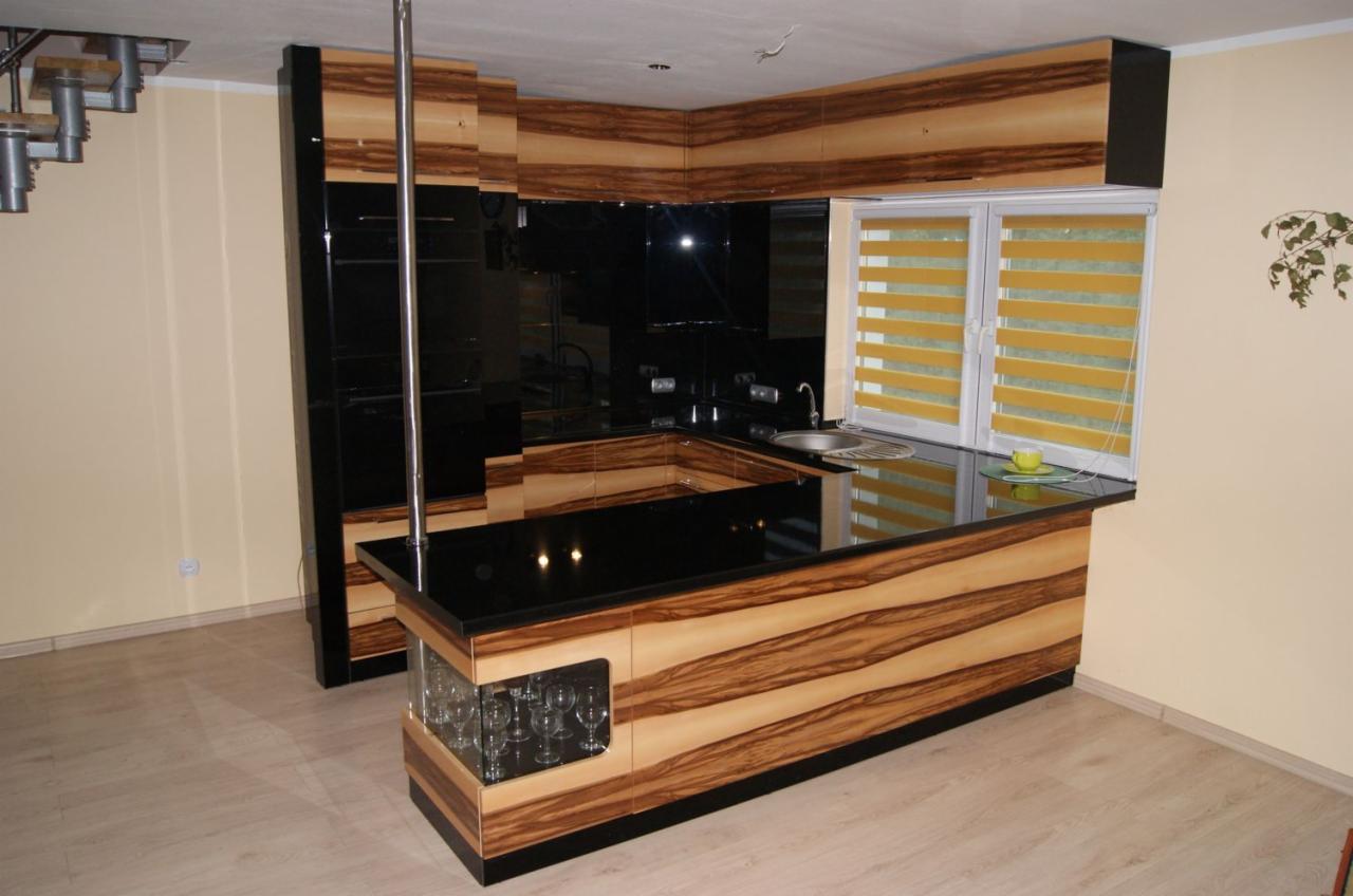 Nowoczesna kuchnia z blatem z czarnego szkła i frontami z egzotycznego drewna zebra. W tle okno z żaluzjami i schody spiralne. Szklana witryna w wyspie.