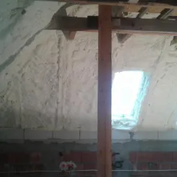 Poddasze z oknami połaciowymi, pokryte warstwą izolacyjną z widoczną strukturą piany, surowa ściana z cegieł w dolnej części, drewniana konstrukcja dachu.