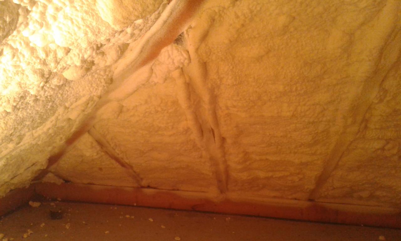 Poddasze ocieplone pianką, widoczne krokwie i nierównomierna struktura piany w ciepłym oświetleniu.