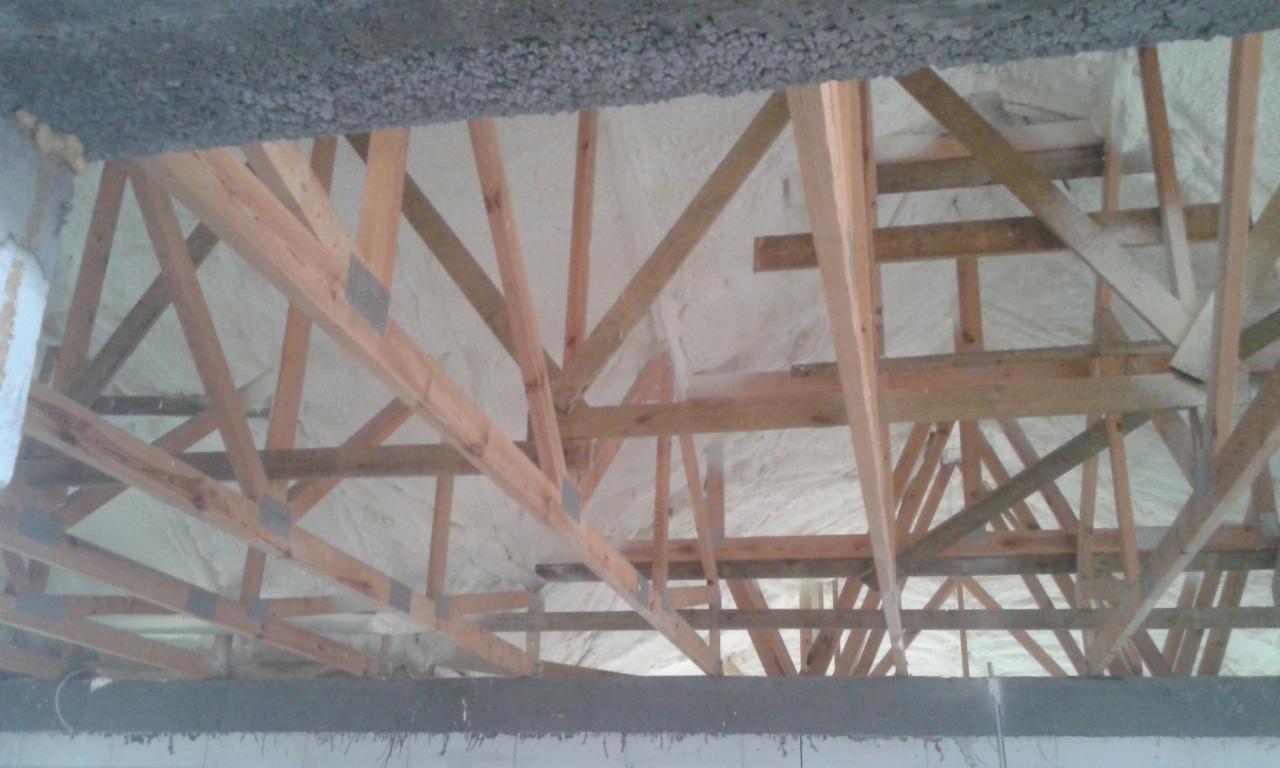 Skośny widok na drewnianą konstrukcję dachu wypełnioną jasną, sztywną pianą izolacyjną, z widocznymi elementami betonowymi u góry i dołu kadru.