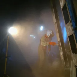 Pracownik w kombinezonie ochronnym i masce natryskuje powłokę na metalową konstrukcję przy użyciu pistoletu lakierniczego, oświetlony reflektorem, z widocznym pyłem w powietrzu.