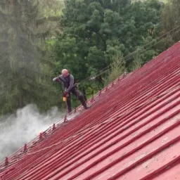 Pracownik w uprzęży zabezpieczającej, czyszczący czerwoną blachodachówkę na stromym dachu za pomocą myjki ciśnieniowej, z widoczną parą wodną i zielonym tłem drzew.