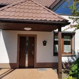 Wejście do domu z brązowymi drzwiami i zadaszeniem z brązowej dachówki, biała elewacja z brązowym cokołem, skrzynka na listy obok drzwi, widoczne okna z firankami.