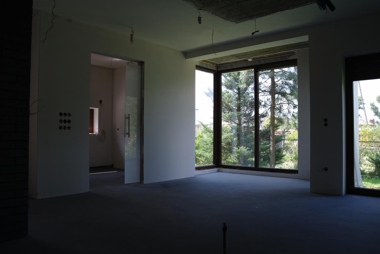 Surowe wnętrze z dużymi oknami i otwartym przejściem. Widoczne instalacje elektryczne, stan deweloperski, sufit bez wykończenia. Duże przeszklenia z widokiem na zieleń.