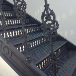 Metalowe schody z ozdobnymi, kutymi balustradami, pomalowane na ciemny kolor. Widoczne ażurowe detale i wz&oacute;r na stopniach. Ujęcie z dołu, ukazujące perspektywę wznoszenia.