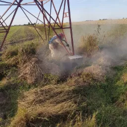 Pracownik w kombinezonie ochronnym i masce piaskuje podstawę metalowej konstrukcji słupa energetycznego na tle pola uprawnego.