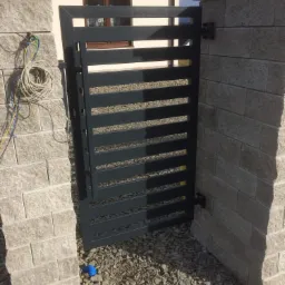 Nowoczesna, stalowa furtka wjazdowa z poziomymi szczeblami, osadzona pomiędzy kamiennymi słupkami. Widoczne okablowanie elektryczne na jednym ze słupków oraz niebieski element na żwirowym podłożu.