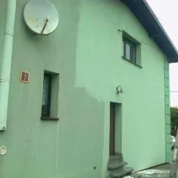 Dom z elewacją w trakcie malowania na zielono, widoczna drabina i częściowo pomalowana ściana, satelita na ścianie. Etap prac malarskich.