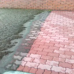 Częściowo wyczyszczona, czerwona kostka brukowa przed ceglanym murem. Lewa strona mokra i brudna, prawa czysta i sucha, ukazująca efekt czyszczenia.