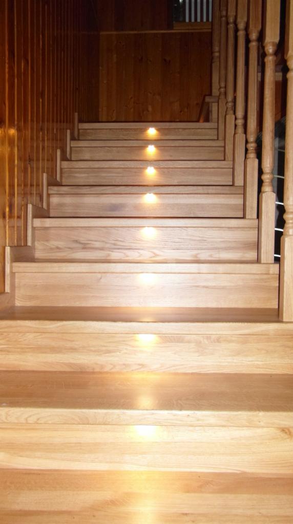 Jasne drewniane schody z oświetleniem punktowym w każdym stopniu, balustrada z toczonymi elementami, wykończenie ścian w drewnie.
