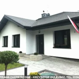 Parterowy dom jednorodzinny z czarną dach&oacute;wką i stolarką okienną, białą elewacją, z flagą Polski zawieszoną przy oknie i zadbanym trawnikiem z ozdobnymi krzewami.