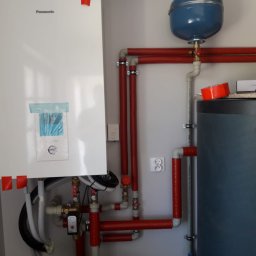 Globeco Adam Nowak - Instalacja kotła grzewczego Panasonic w Krakowie z czerwonymi izolacjami na rurach, widoczny zbiornik wyrównawczy i fragment instalatora.