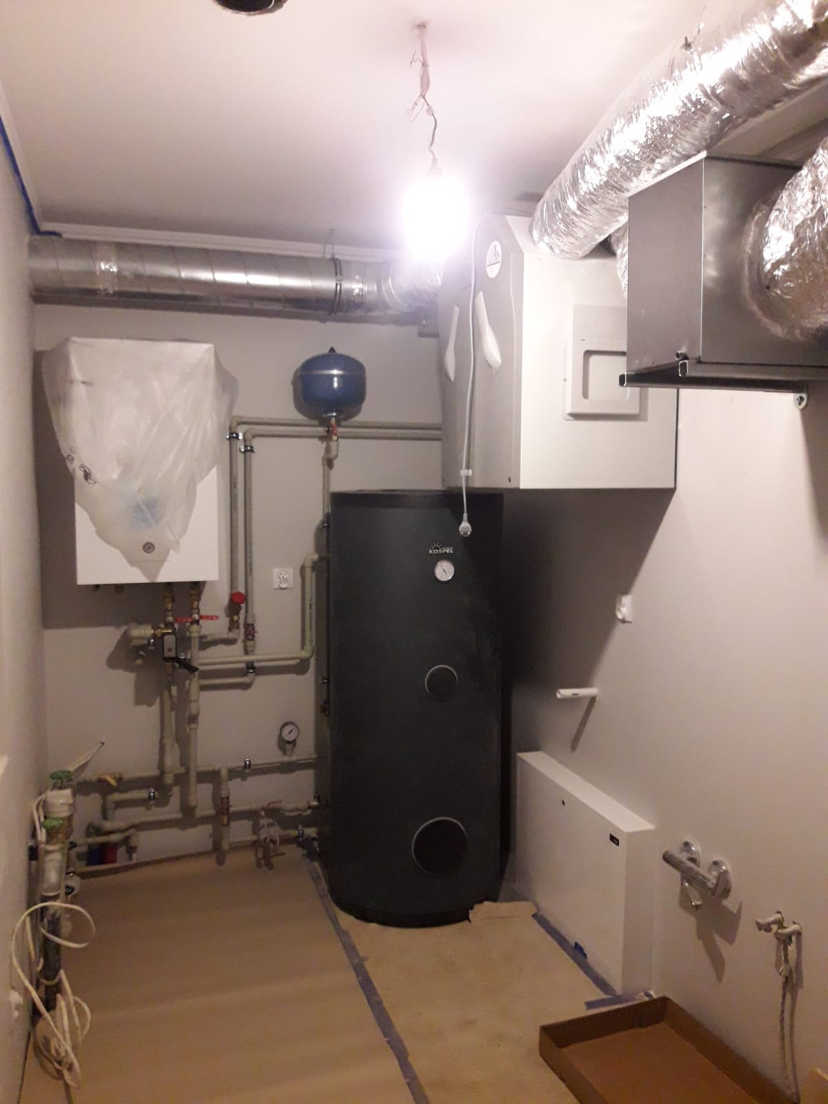 Wnętrze kotłowni w trakcie instalacji, widoczny zbiornik akumulacyjny Kospel, biały zasobnik ciepłej wody osłonięty folią, rury instalacyjne, kanały wentylacyjne i elementy rekuperacji pod sufitem,...