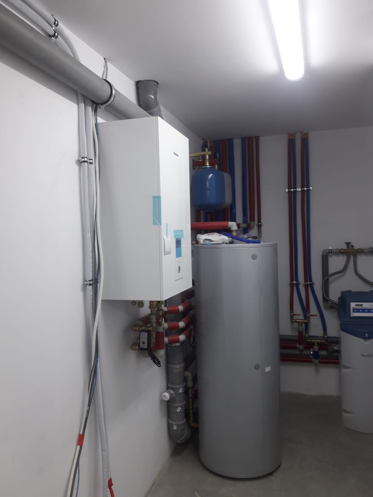 Kompletna instalacja grzewcza w kotłowni: biały piec, srebrny zbiornik, niebieskie naczynie przeponowe, liczne rury miedziane i plastikowe w kolorach czerwonym i niebieskim, filtr wody na ścianie...