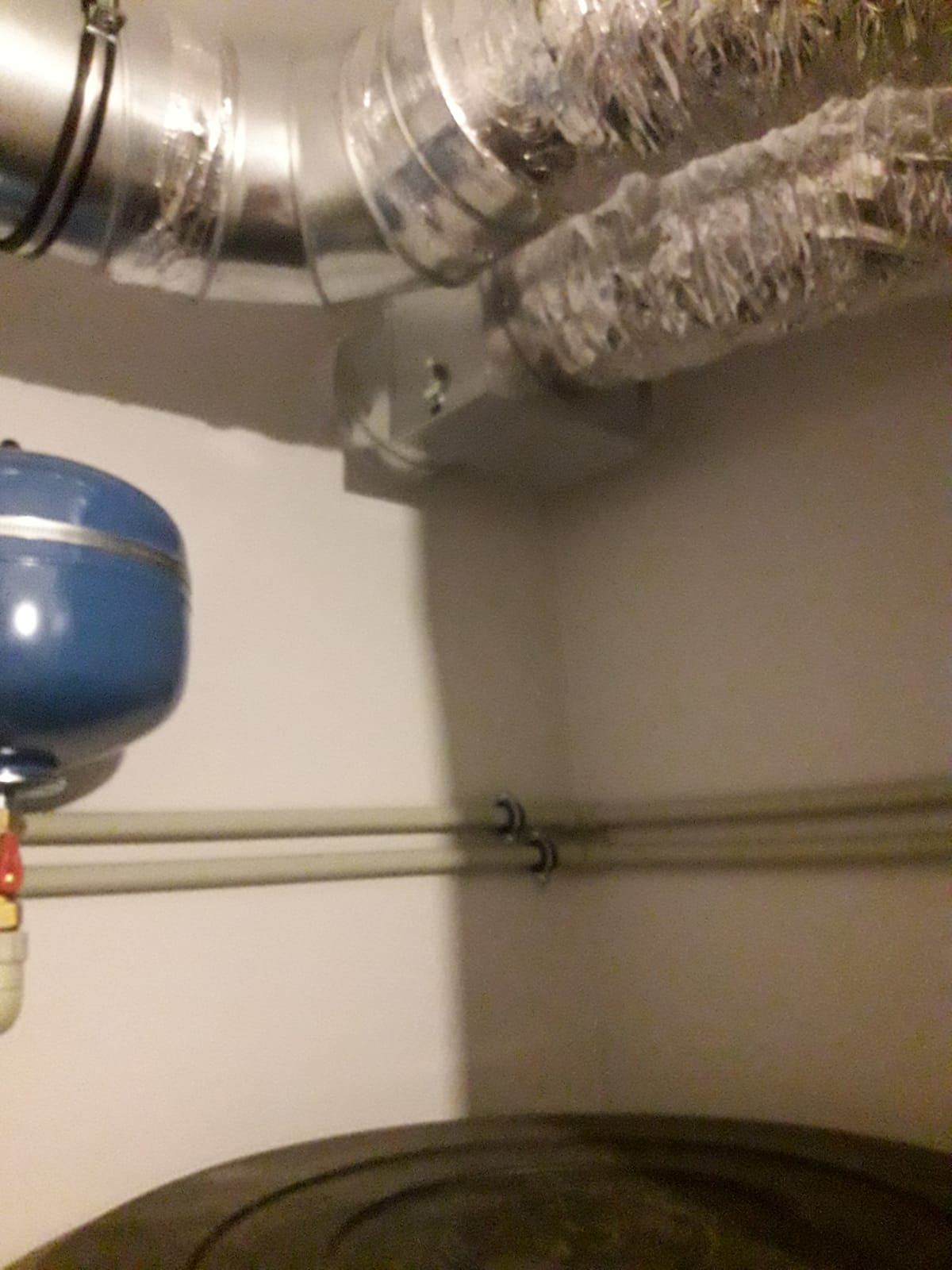 Fragment instalacji z widocznymi rurami wentylacyjnymi owiniętymi folią izolacyjną, zbiornikiem ciśnieniowym w kolorze niebieskim oraz rurami instalacji grzewczej przymocowanymi do ściany białymi...