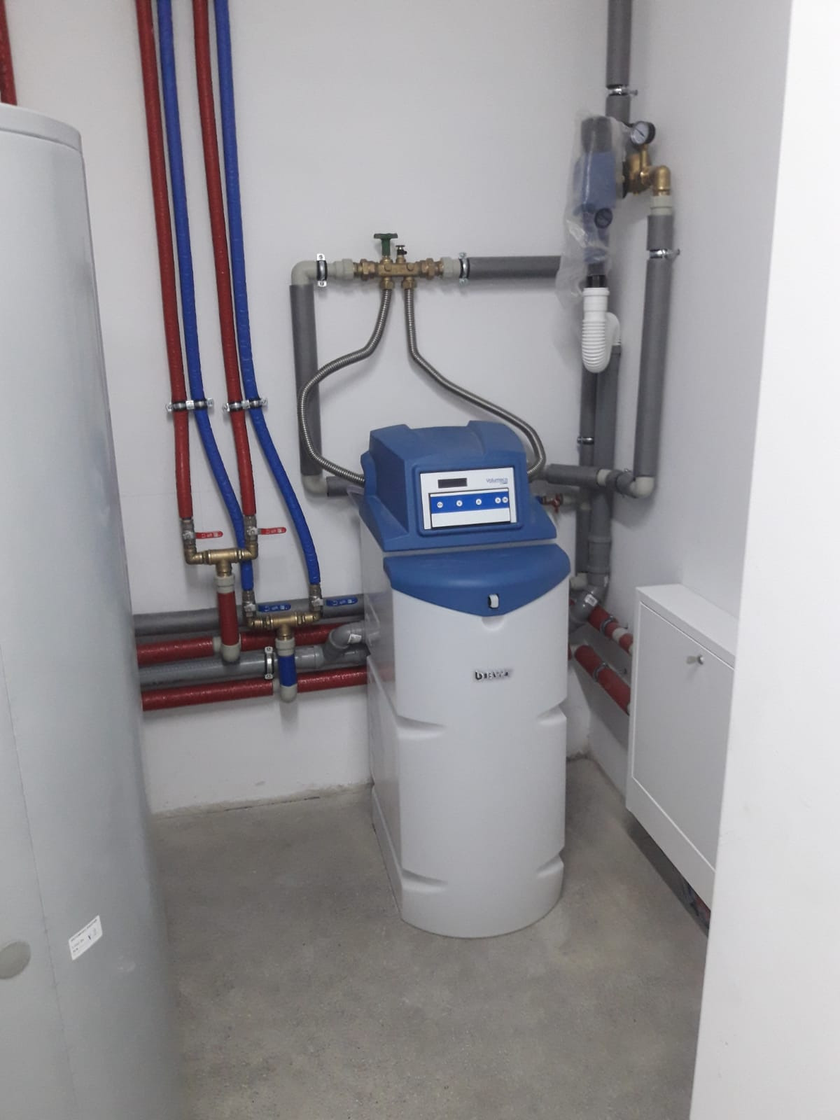 Biały zmiękczacz wody BWT z niebieską górną częścią, podłączony do instalacji wodnej z rurami czerwonymi, niebieskimi i szarymi w pomieszczeniu technicznym z szarą podłogą.