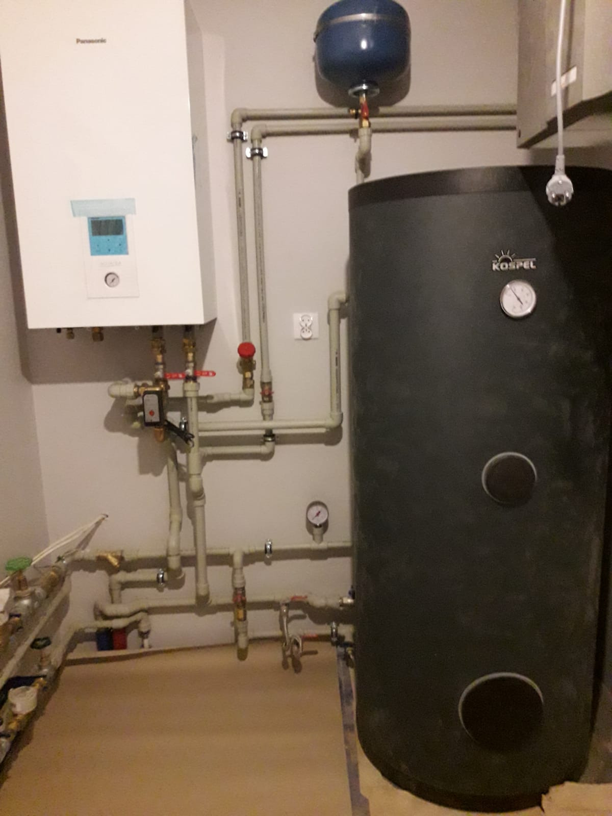 Zestaw urządzeń grzewczych: biały piec Panasonic, pionowy czarny zasobnik Kospel z manometrem, niebieski zbiornik wyrównawczy, połączone rurami z zaworami w pomieszczeniu technicznym.