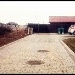 Szeroki podjazd z jasnej kostki brukowej, obrzeża z czerwonej kostki. W tle dom z czerwonym dachem, ogrodzenie i zaparkowany samochód.