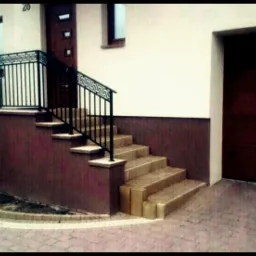 Zewnętrzne schody z jasnych płytek prowadzą do drzwi domu. Czarna metalowa balustrada z greckim wzorem. Obok garaż z brązową bramą. Kostka brukowa na podjeździe.