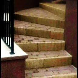 Schody zewnętrzne wyłożone kostką brukową w odcieniach beżu i czerwieni, z czarną metalową balustradą, prowadzące do góry. Widok z dołu, oświetlenie naturalne.