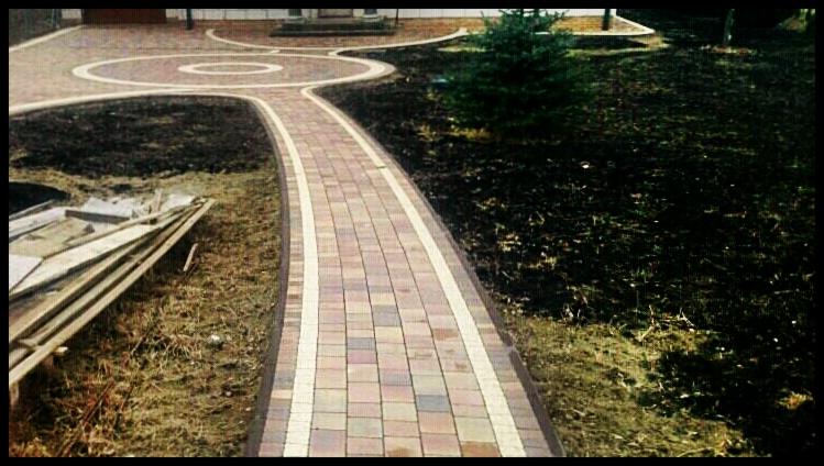 Ogród z ścieżką z kostki brukowej w odcieniach czerwieni i szarości, obrzeżoną białymi pasami. Widoczne rabaty i młode drzewko. Na pierwszym planie deski.