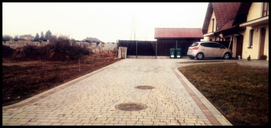 Szeroki podjazd z jasnej kostki brukowej, obrzeża z czerwonej kostki. W tle dom z czerwonym dachem, ogrodzenie i zaparkowany samochód.