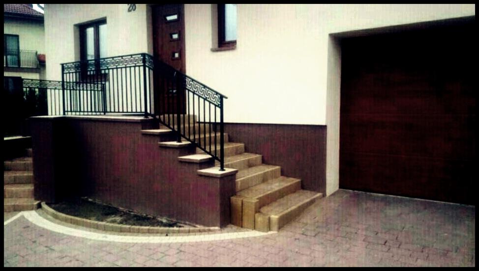 Zewnętrzne schody z jasnych płytek prowadzą do drzwi domu. Czarna metalowa balustrada z greckim wzorem. Obok garaż z brązową bramą. Kostka brukowa na podjeździe.