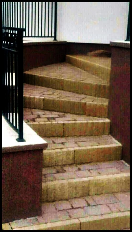 Schody zewnętrzne wyłożone kostką brukową w odcieniach beżu i czerwieni, z czarną metalową balustradą, prowadzące do góry. Widok z dołu, oświetlenie naturalne.