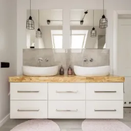 Nowoczesna łazienka z dwiema owalnymi umywalkami nablatowymi, blatem z płyty OSB, białymi szafkami z podłużnymi uchwytami, dwoma lustrami, industrialnym oświetleniem i różowymi dywanikami.