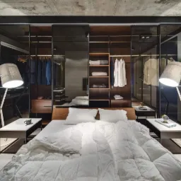 Nowoczesna sypialnia z łóżkiem, szafkami nocnymi i dużą garderobą za przesuwnymi, szklanymi drzwiami, oświetloną przez dwie lampy podłogowe z abażurami w kolorze szarym.