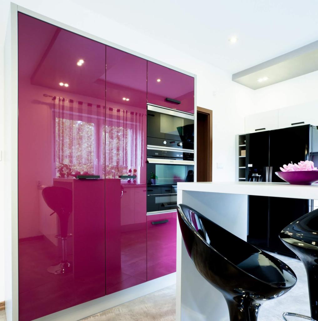 Nowoczesna kuchnia z frontami szafek w kolorze fuksji o wysokim połysku, zintegrowanymi piekarnikami i czarnymi lodówkami, widoczne krzesła barowe i elementy wystroju.
