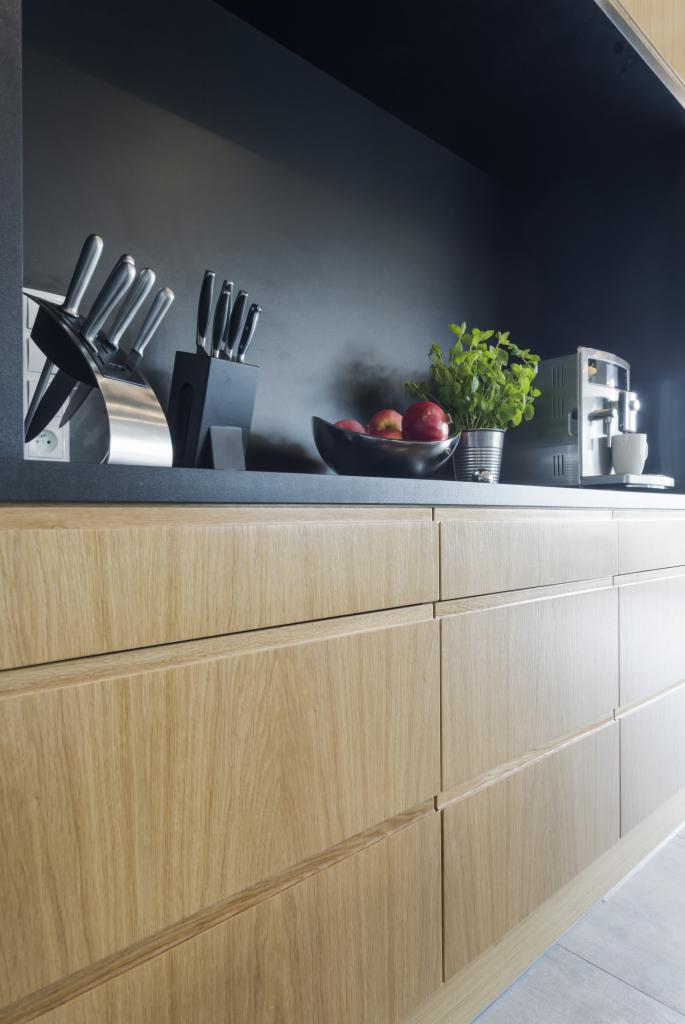 Fronty szafek kuchennych z jasnego drewna, na blacie zestaw noży, misa z jabłkami, zioła w doniczce i ekspres do kawy.