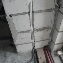 Białe przewody elektryczne prowadzone w bruździe ściany z betonu komórkowego, zabezpieczone peszlem przy podłodze. Szare tło surowego betonu.