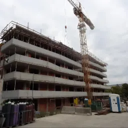 Wielopiętrowy budynek w stanie surowym otwartym, widoczny żuraw budowlany, kontenery z butlami gazowymi i przenośna toaleta na placu budowy.