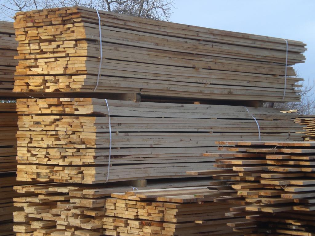 Stosy jasnego drewna budowlanego, ułożone na paletach i zabezpieczone białym sznurkiem. Widok na składowane deski na zewnątrz.