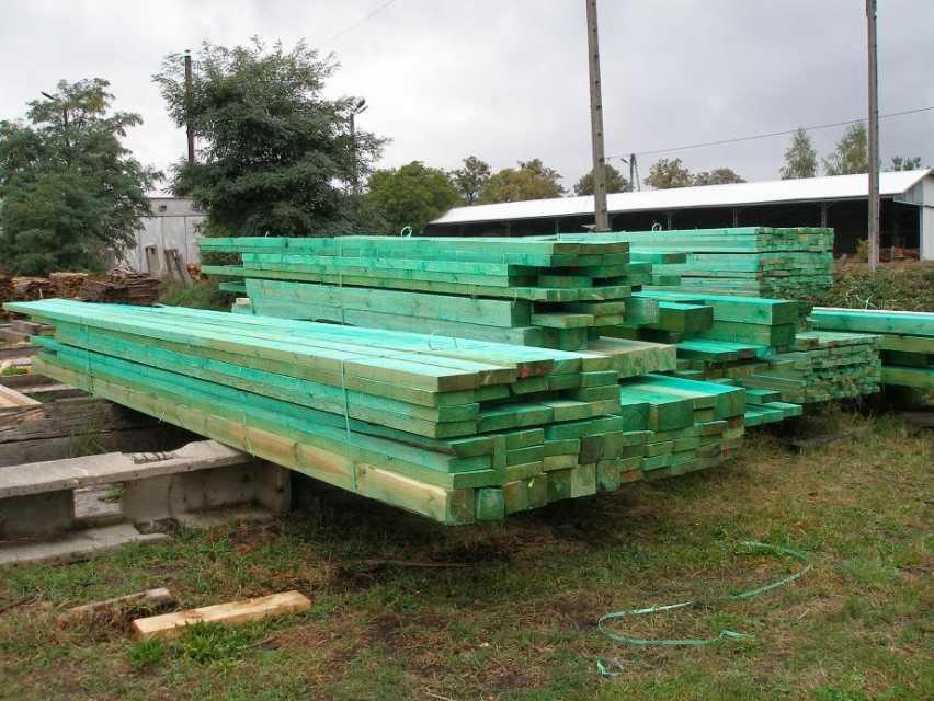 Stosy impregnowanego na zielono drewna budowlanego, ułożone na zewnątrz, na tle zieleni i budynku magazynowego z białym dachem, w pochmurny dzień.