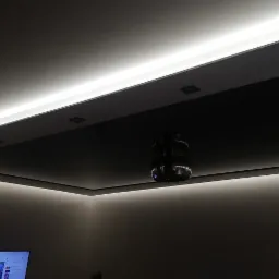 Nowoczesny sufit podwieszany z oświetleniem LED, centralnie umieszczony projektor, minimalistyczny design, ciemne elementy kontrastujące z jasnym światłem.