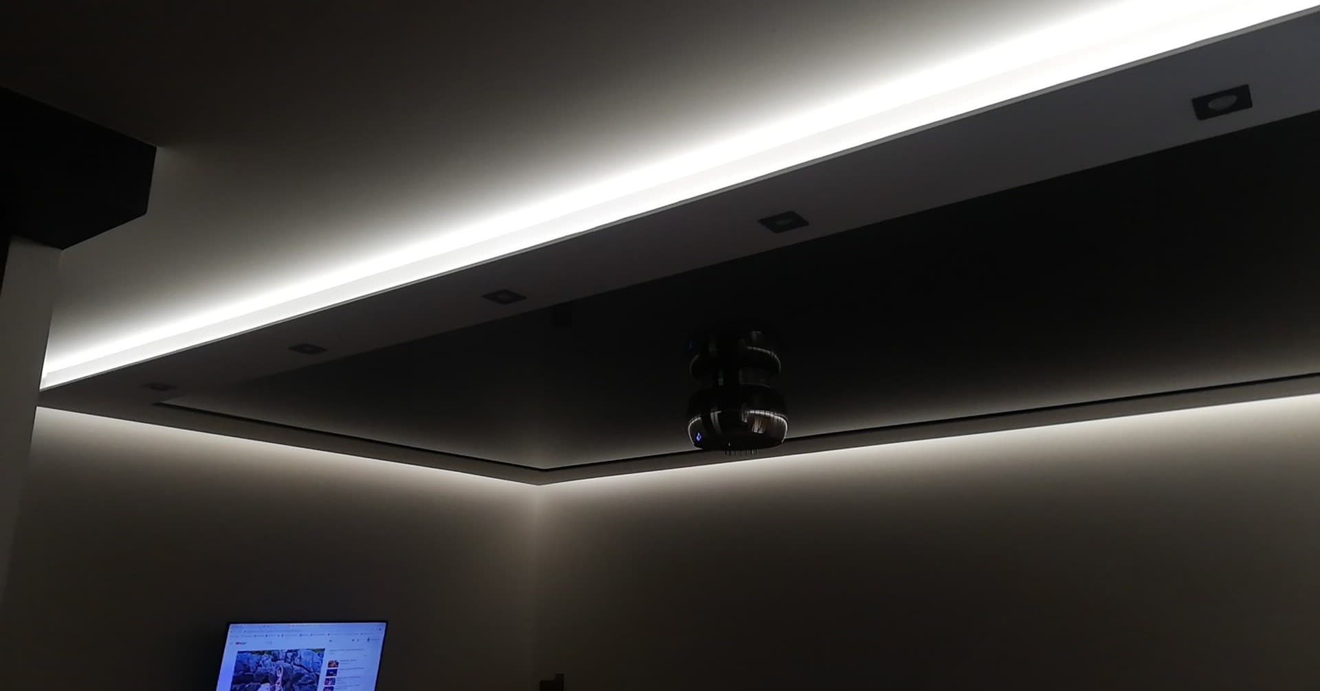 Nowoczesny sufit podwieszany z oświetleniem LED, centralnie umieszczony projektor, minimalistyczny design, ciemne elementy kontrastujące z jasnym światłem.