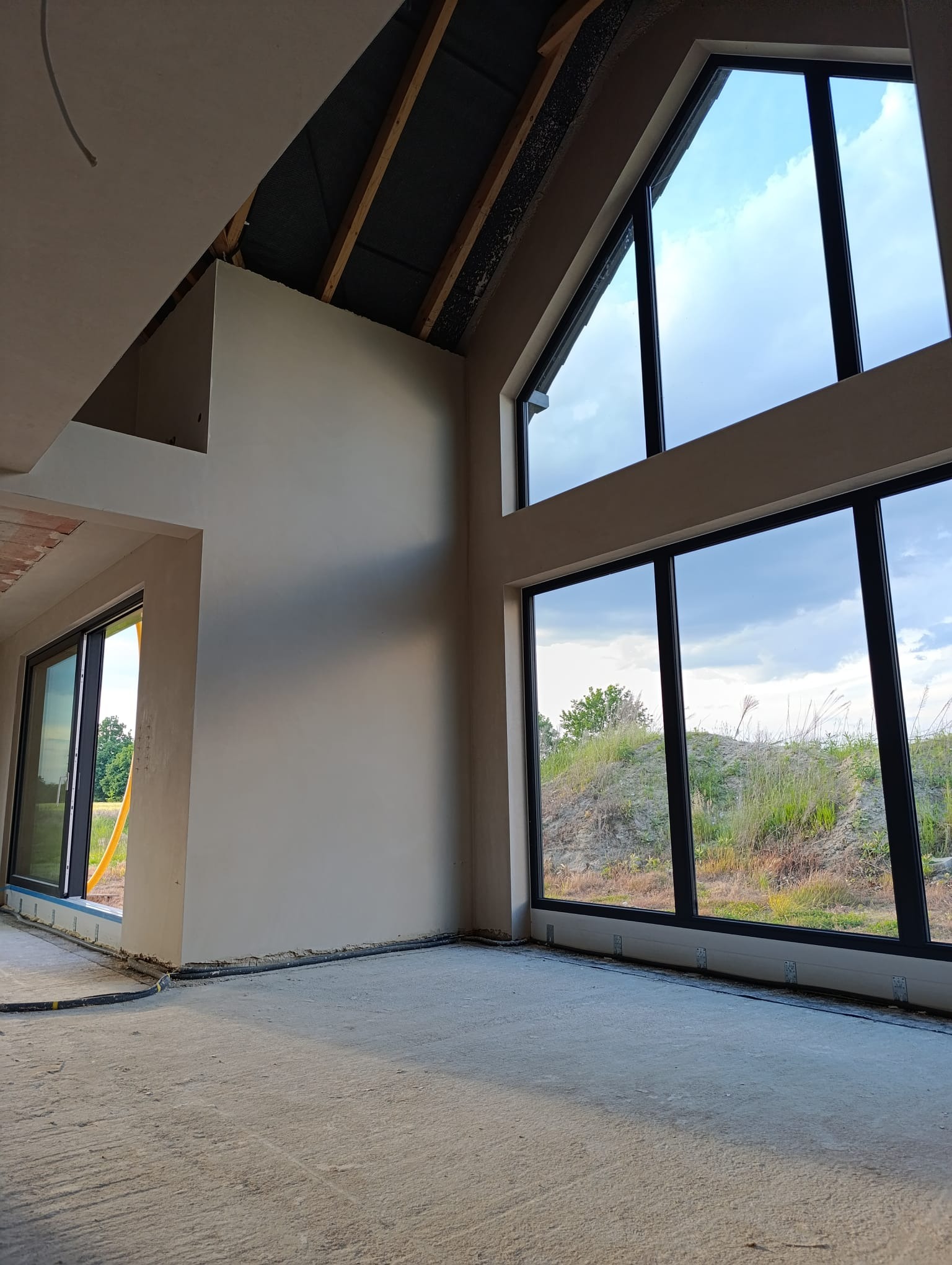 Wnętrze domu w trakcie wykańczania: gładkie, jasne ściany po tynkowaniu, duże okna z widokiem na zewnątrz, surowa podłoga. Widoczne elementy konstrukcji dachu.