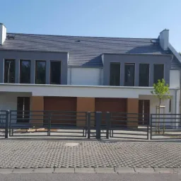 Nowoczesny bliźniak z szarą elewacją i ciemnymi oknami, widok z zewnątrz przez metalowe ogrodzenie, kostka brukowa na podjeździe.