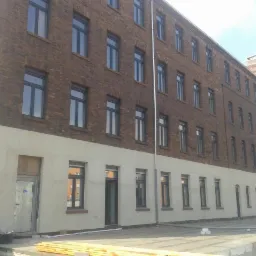 Wysoki budynek z cegły z nowymi, ciemnymi oknami i świeżo otynkowaną dolną partią elewacji. Widoczne drewniane elementy konstrukcyjne i fragment osoby w lewym dolnym rogu.