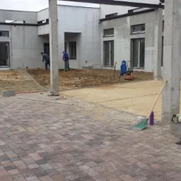 Układanie kostki brukowej na posesji, widoczny fragment ułożonej nawierzchni z kostki, w tle pracownicy przygotowują podłoże pod dalszą część prac.