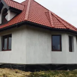 Wykończony dom z czerwoną dachówką i jasną elewacją. Widoczne okna z brązowymi ramami, fragment fundamentu z czarną folią. Ujęcie z boku, w tle pochmurne niebo.