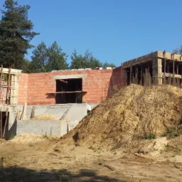 Surowy stan budynku z czerwonej cegły, z widocznym zbrojeniem i drewnianymi szalunkami, otoczony ziemią i drzewami w słoneczny dzień. Etap wznoszenia budynku.