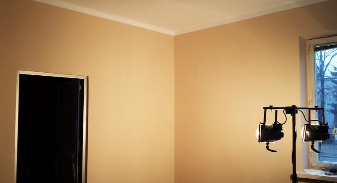 Ściana w odcieniach beżu po malowaniu, widoczna listwa przysufitowa, drzwi z czarnym wnętrzem po lewej, okno z widokiem na zewnątrz i lampa oświetleniowa po prawej.