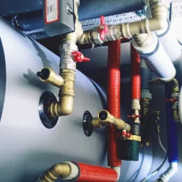 Szczegółowe ujęcie instalacji hydraulicznej z zaworami, rurami miedzianymi i plastikowymi, pokrytymi izolacją termiczną, zamontowanej na zbiorniku z logo firmy TopInstall.